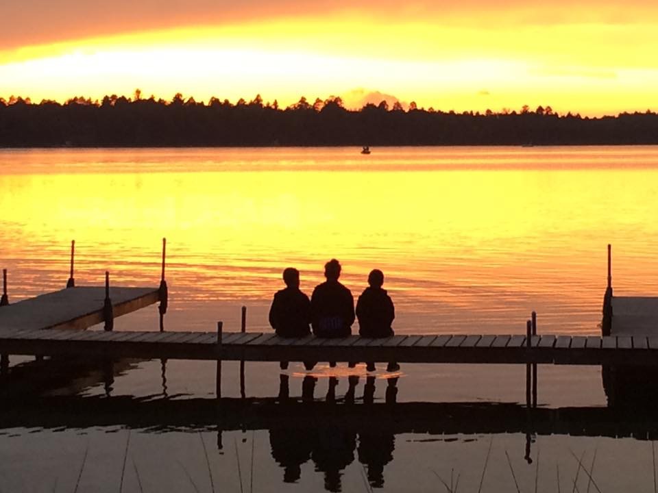 Three people sitting on dock at sunset.